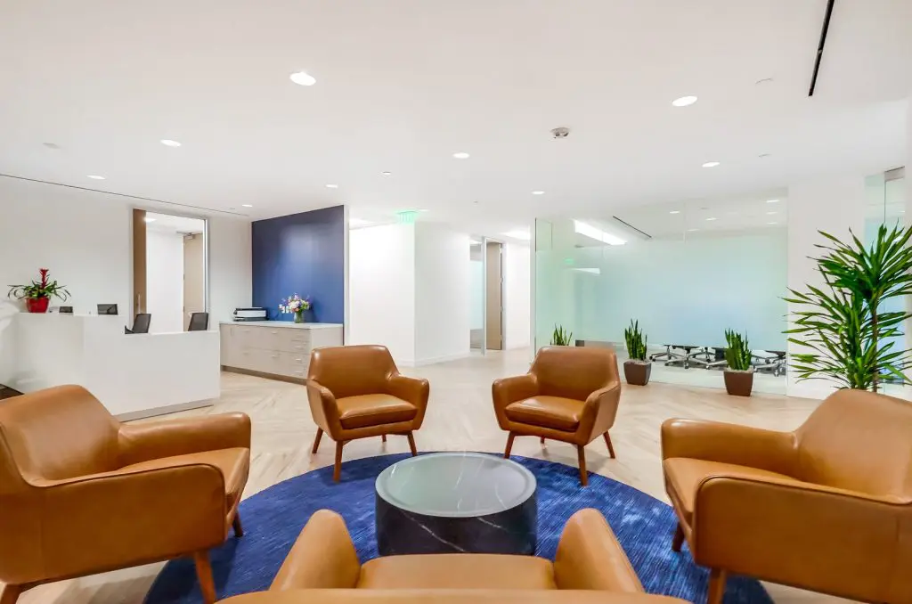 Barrister Suites Office Lease Modern office lobby with four brown leather chairs arranged in a circle around a glass coffee table on a blue rug, plants in the background, and white walls with bright lighting.