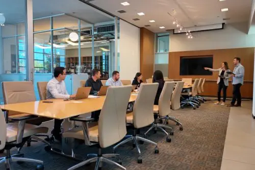 Barrister Suites Office Lease A group of people sit at a long conference table in a modern office, while two people stand near a large screen, with one pointing toward it as if presenting. The room has glass walls and office chairs.