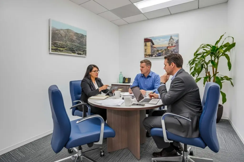 Barrister Suites Office Lease Three people in business attire sit around a round table in a modern office, discussing documents and using laptops. There are pictures on the wall and a large plant in the corner.