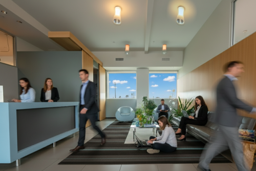 Barrister Suites Office Lease Modern office lobby with people in business attire; some are sitting on couches and chairs working on laptops, while others stand or walk. Large windows show a blue sky outside. Reception desk is on the left.