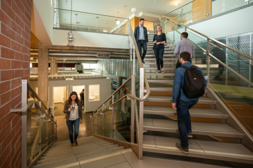 Barrister Suites Office Lease People walk up and down a modern indoor staircase in a bright building. Some are using their phones or talking, and others carry backpacks. The space features glass walls, metal railings, and natural light.