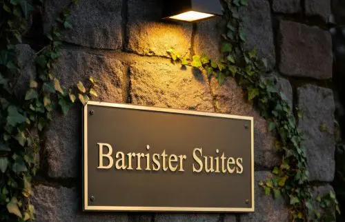 Barrister Suites Office Lease A brass sign reading Barrister Suites is mounted on a stone wall with ivy and illuminated by a light above. A black metal fence is visible in the foreground.