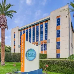 Barrister Suites Office Lease A modern office building with orange and beige panels, surrounded by palm trees and manicured shrubs. A sign in the foreground reads 1600 Rosecrans Avenue, MBS Media Campus.