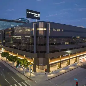 Barrister Suites Office Lease A modern, multi-story office building at a city intersection, illuminated at dusk, with City National Bank signage on the rooftop and glass windows reflecting the evening sky.