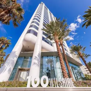 Barrister Suites Office Lease A tall white office building with curved corners and large glass windows is surrounded by palm trees. The address 100 Wilshire is displayed in large letters in front. The sky is blue with scattered clouds.
