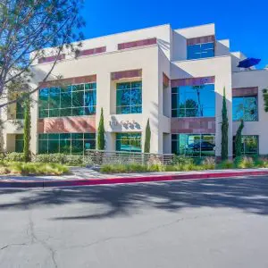 Barrister Suites Office Lease A modern three-story office building with large reflective windows, surrounded by trees and landscaping, under a clear blue sky. The building sits beside a wide, paved road with a red curb.