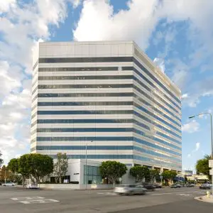 Barrister Suites Office Lease A modern, white mid-rise office building with reflective glass windows stands on a busy street corner, surrounded by trees, cars, and people under a partly cloudy sky.