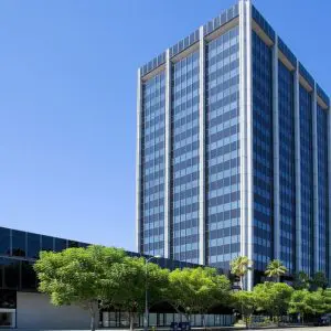 Barrister Suites Office Lease A tall modern office building with reflective blue-tinted windows stands beside a shorter building, surrounded by green trees and set against a clear blue sky.