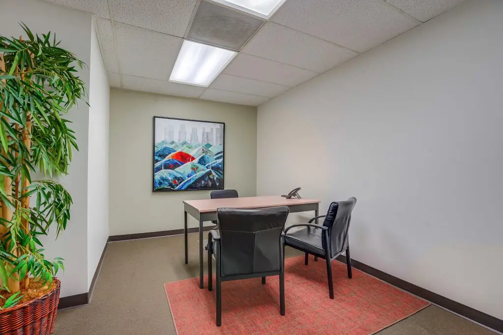 Barrister Suites Office Lease A small office with a desk, two black chairs, a phone, a potted plant, a red rug, and a colorful painting of hills on the wall. The space has white walls and ceiling lights.