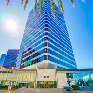 Barrister Suites Office Lease A modern glass high-rise building with 1801 and Preferred Bank at the entrance, viewed from below on a sunny day with palm tree leaves and a bright sun in the blue sky above.