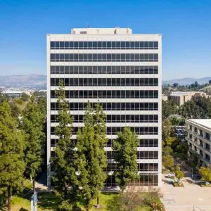 Barrister Suites Office Lease A tall office building with many windows stands behind several tall green trees, next to another office building under a clear blue sky. Some parked cars and distant hills are visible in the background.