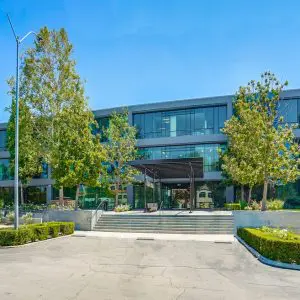 Barrister Suites Office Lease A modern three-story office building with large glass windows, surrounded by trees and landscaping, under a clear blue sky. A driveway and walkway lead to the main entrance with steps and a metal canopy.