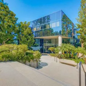 Barrister Suites Office Lease A modern glass office building surrounded by lush green trees and landscaped plants, with a concrete pathway and railings leading to the entrance under a clear blue sky.