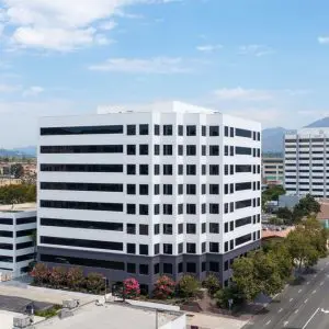 Barrister Suites Office Lease A modern, mid-rise office building with white and black horizontal stripes stands beside a wide, empty street lined with trees. Other office buildings and mountains are visible in the background under a partly cloudy sky.