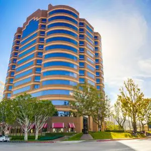 Barrister Suites Office Lease A modern, multi-story office building with curved edges and many windows, surrounded by trees and a clear blue sky. The sun is shining brightly, casting shadows on the street and sidewalk in front of the building.