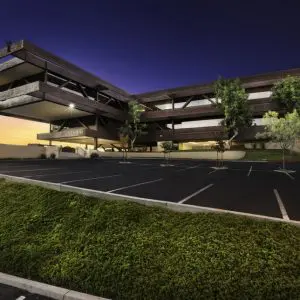Barrister Suites Office Lease A modern, angular office building with large overhangs and glass windows is shown at dusk, with lights on inside. The parking lot in front is empty, and there are trees and green landscaping in the foreground.