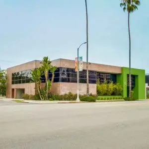 Barrister Suites Office Lease A modern, two-story building with large glass windows and a green accent wall sits on a street corner lined with palm trees and banners. The street and sidewalks appear empty.