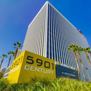 Barrister Suites Office Lease Tall modern office building with vertical lines, surrounded by palm trees and greenery. A large yellow sign in front reads 5901 Century. The sky is clear and blue.