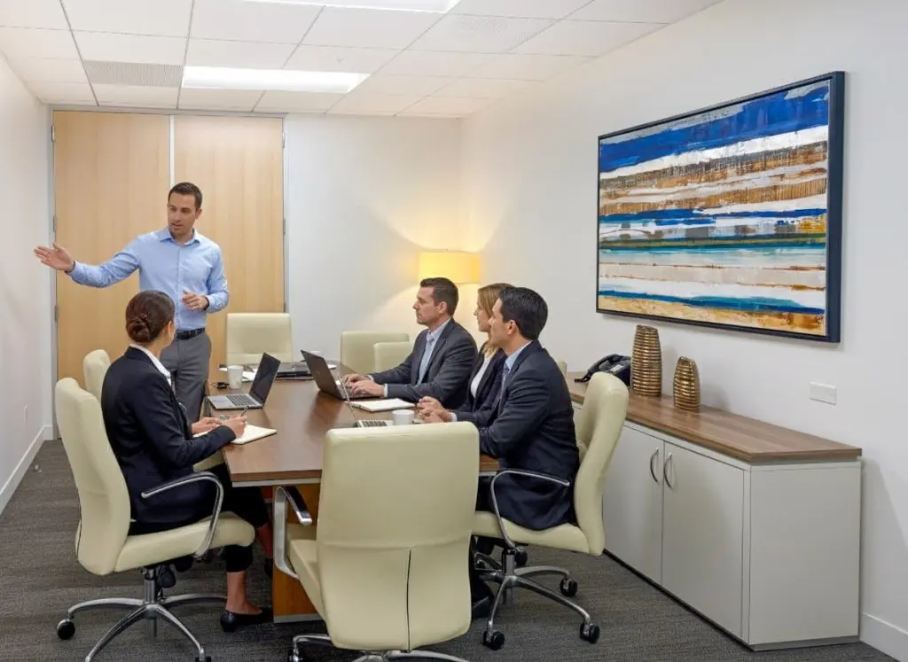 Barrister Suites Office Lease A man stands and gestures while speaking to three colleagues seated at a conference table in a modern office with laptops; a colorful abstract painting hangs on the wall.