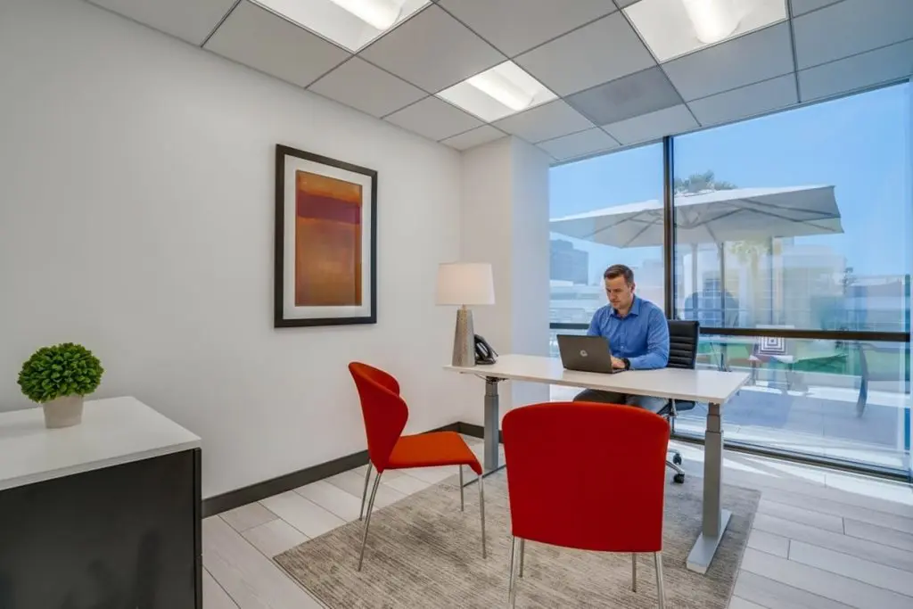 Barrister Suites Office Lease A man sits at a desk working on a laptop in a modern office with large windows, red chairs, a potted plant, framed artwork, and an outdoor patio visible through the window.