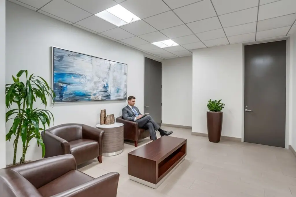 Barrister Suites Office Lease A man in a suit sits on a brown leather chair in a modern office waiting area with another chair, coffee table, two potted plants, and an abstract painting on the wall. The space has white walls and a tiled floor.