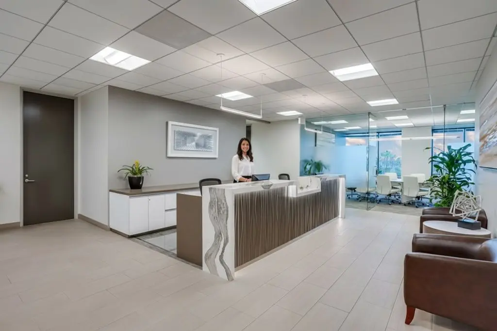 Barrister Suites Office Lease A woman stands behind a modern reception desk in a bright, spacious office lobby with large windows, potted plants, and a conference room visible in the background.