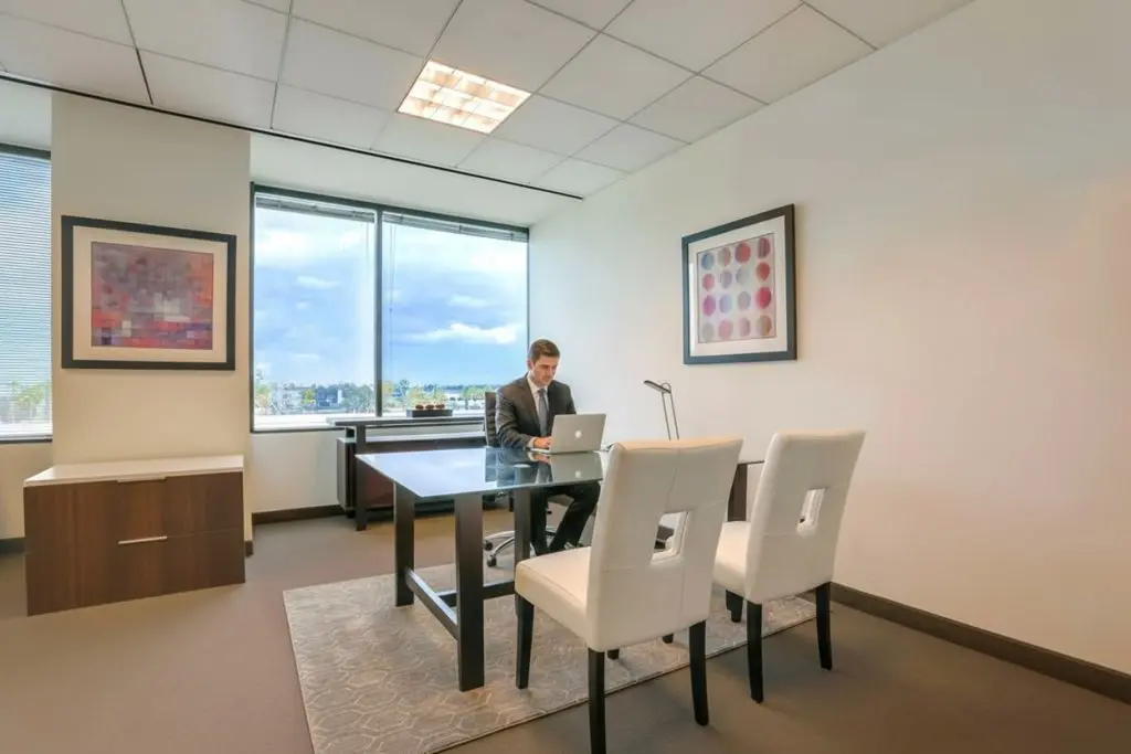 Barrister Suites Office Lease A man in a suit works on a laptop at a modern office desk. The office has large windows, two white chairs facing the desk, abstract art on the walls, and neutral-colored furnishings.