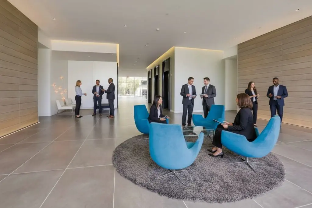 Barrister Suites Office Lease A modern office lobby with groups of professionally dressed people standing and talking, and four people seated around a glass table in bright blue chairs on a gray rug. The space is spacious and well-lit.