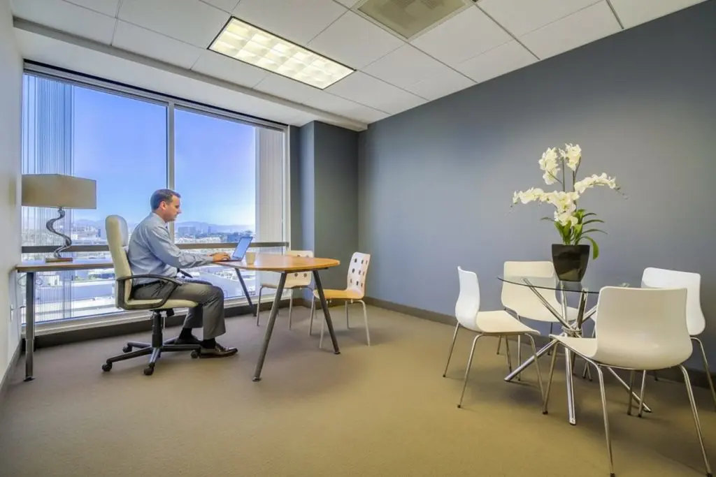 Barrister Suites Office Lease A man works on a laptop at a wooden desk in a modern office with large windows, gray walls, a round glass table with white chairs, and a potted white orchid.