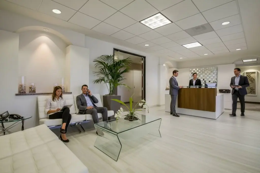 Barrister Suites Office Lease Modern office lobby with a glass coffee table, white seating, and potted plants. Two people wait on chairs, while three others interact at the reception desk. The space is bright, with white walls and tile flooring.