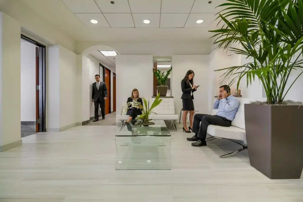Barrister Suites Office Lease Four people in business attire are in a modern office waiting area with white walls, glass table, green plants, and white chairs. Two sit using phones, one sits with a laptop, and one stands near the hallway entrance.