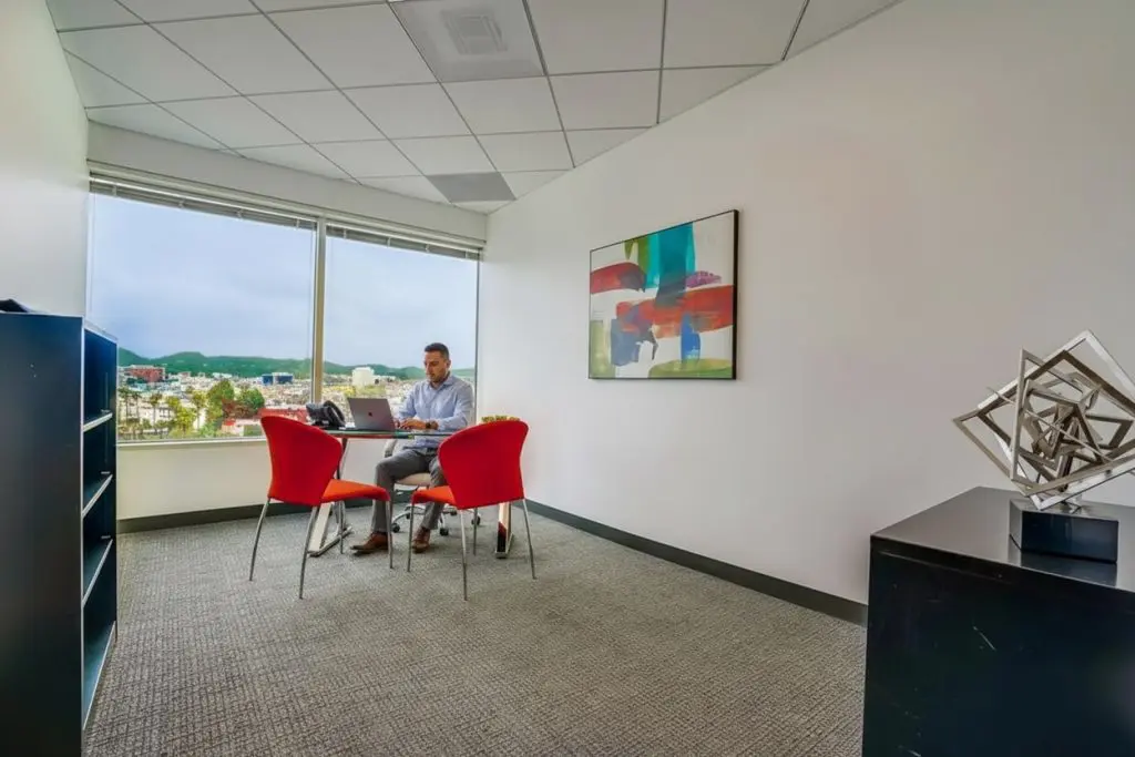 Barrister Suites Office Lease A man works on a laptop at a small desk with two red chairs in a modern office with large windows, abstract wall art, and geometric sculpture on a cabinet.
