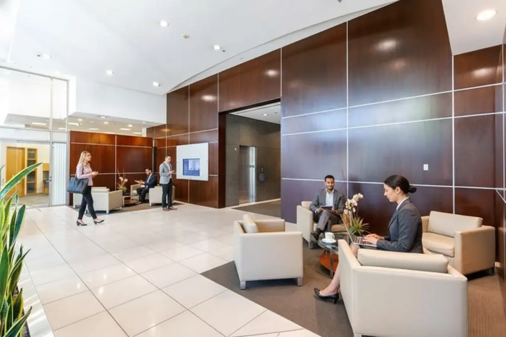 Barrister Suites Office Lease Modern office lobby with wood-paneled walls and white tile floors. Several people are seated and talking, while others are walking or engaging in conversation. Plants and a wall screen decorate the space.