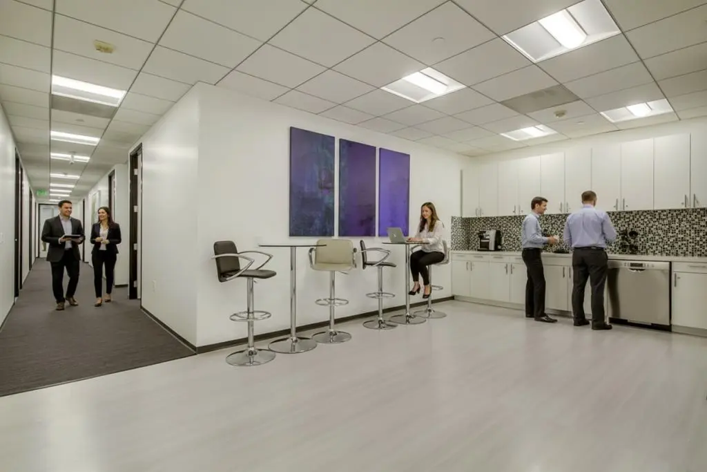 Barrister Suites Office Lease Modern office kitchen with white cabinets, bar stools, and mosaic backsplash. Three people stand by the counter; one person sits working on a laptop. Two people walk down the adjacent hallway. Large abstract art on the wall.