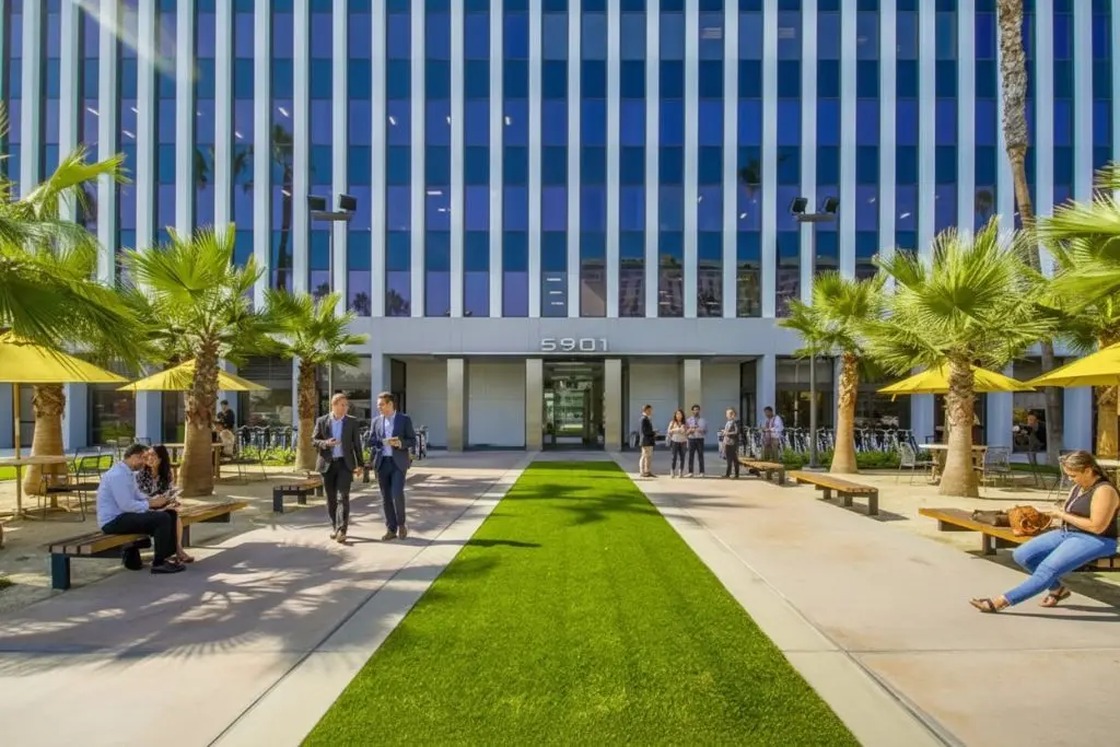 Barrister Suites Office Lease People sit and walk along a pathway with artificial grass, surrounded by palm trees and benches, in front of a modern office building with vertical blue and white stripes and the address number 5901 above the entrance.