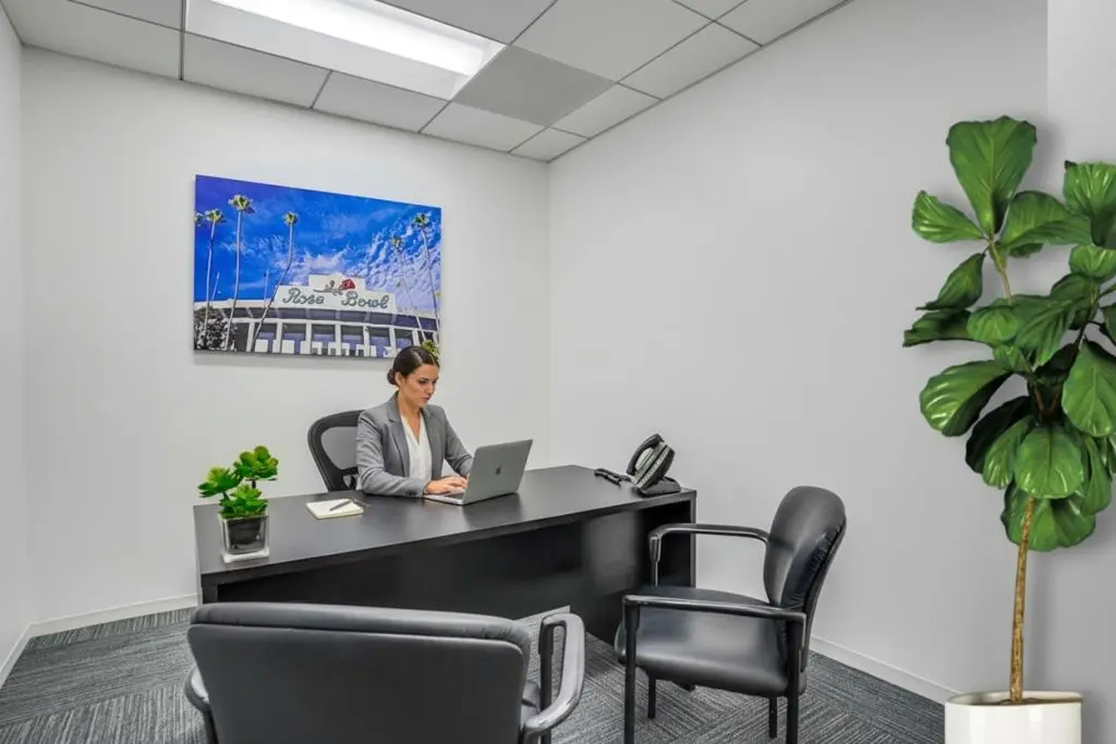 Barrister Suites Office Lease A woman works on a laptop at a black desk in a small, modern office with white walls, a potted plant, two guest chairs, and a large photo of the Rose Bowl hanging on the wall.