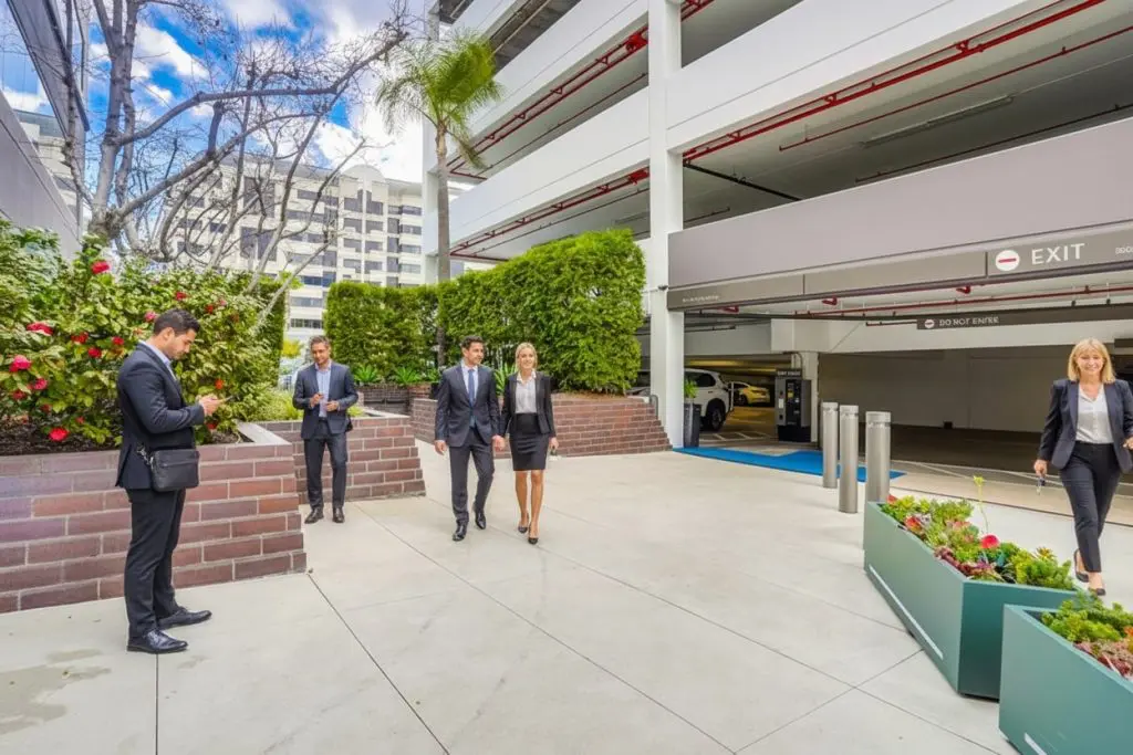 Barrister Suites Office Lease Business professionals walk and talk outside a modern parking garage, with some using phones and others conversing. The area features greenery, planters, and tall buildings in the background under a partly cloudy sky.