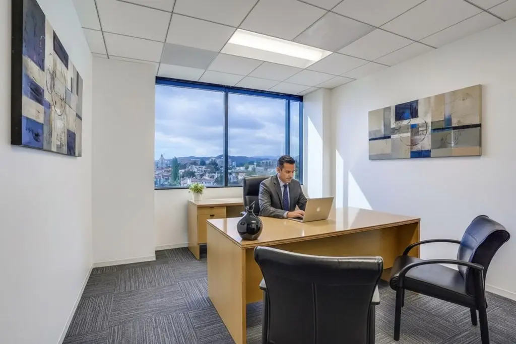 Barrister Suites Office Lease A man in a suit works on a laptop at a wooden desk in a modern office with large window, abstract wall art, and black chairs. Cityscape and cloudy sky visible outside.
