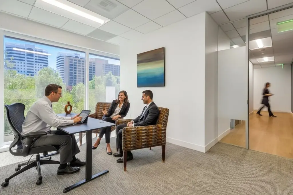 Barrister Suites Office Lease Three people in business attire have a meeting in a modern office with large windows and city views. Two sit across from one at a desk, while another person walks by in the hallway outside a glass door.