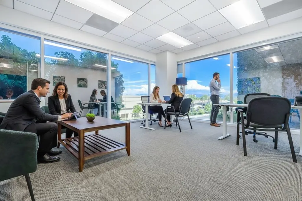 Barrister Suites Office Lease Modern office space with large windows; two people in suits converse at a coffee table, two women work together at a desk, and a man stands near the window talking on a phone. Natural light fills the room.