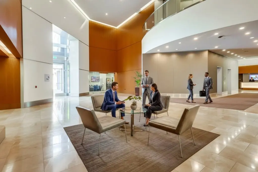 Barrister Suites Office Lease Business professionals in formal attire meet and talk in a modern office lobby with sleek furniture, marble floors, and high ceilings. Other people walk and work in the background, highlighting a corporate environment.