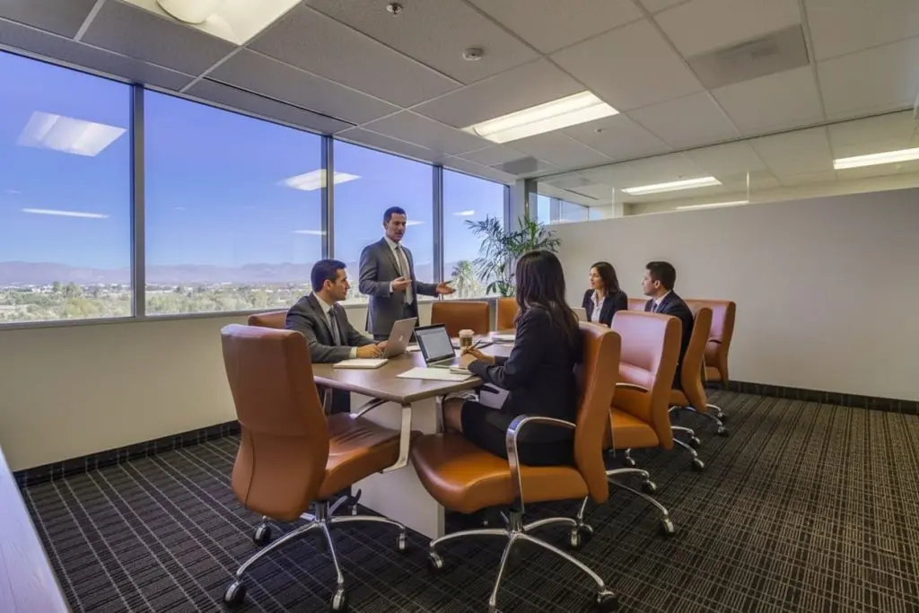 Barrister Suites Office Lease Five people in business attire have a meeting in a modern office with large windows and brown chairs. One person is standing and speaking, while the others sit around a table with laptops and papers.