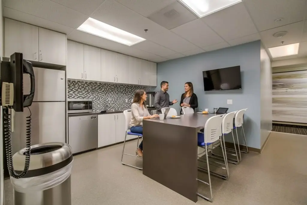 Barrister Suites Office Lease Three people talk in a modern office kitchen, with two seated at a counter and one standing. The room has white cabinets, a microwave, a fridge, and a wall-mounted TV. A trash can and phone are in the foreground.