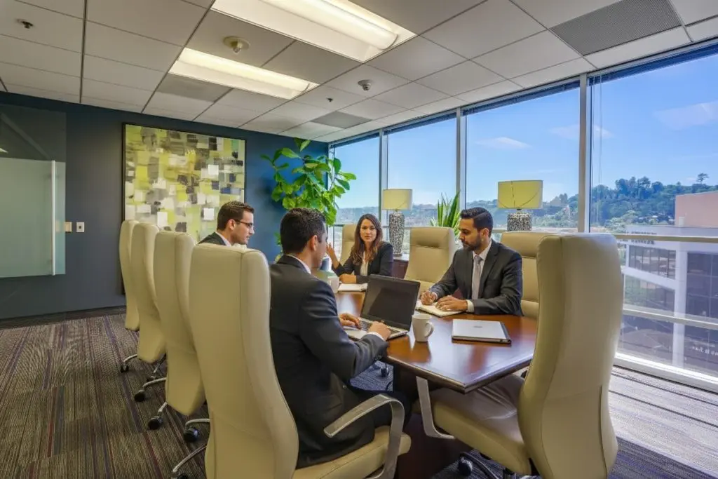 Barrister Suites Office Lease Four business professionals sit around a conference table in a modern office, discussing work. Large windows provide natural light and a scenic outdoor view, while office decor includes artwork and a potted plant.