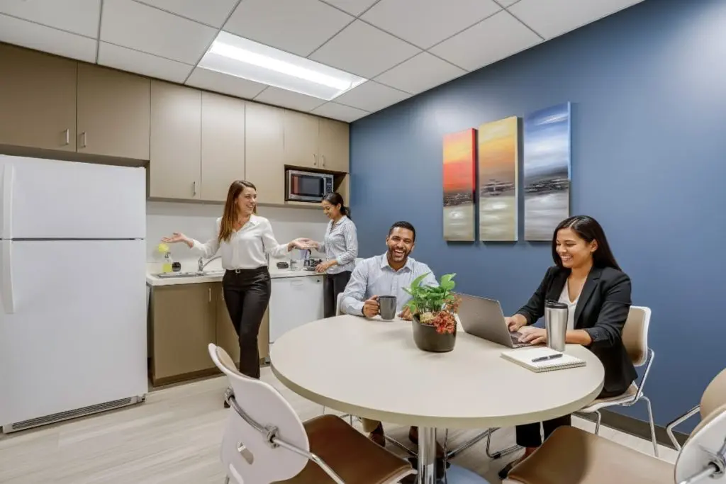 Barrister Suites Office Lease Four colleagues are socializing in a modern office kitchen. Two women stand by the counter, one gesturing while the other makes coffee. A man and a woman sit at a round table, smiling and working on a laptop.
