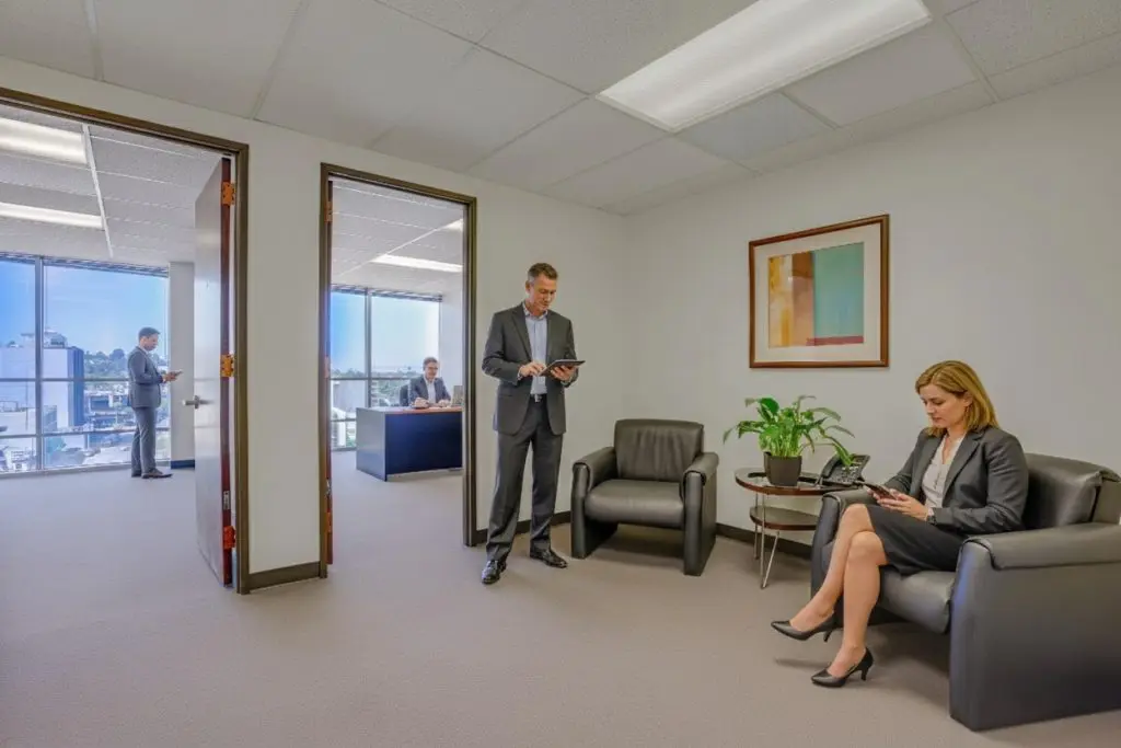 Barrister Suites Office Lease Four people in business attire are in a modern office suite. One woman sits on a black chair using her phone, a man stands using a tablet, and two men are in a glass-walled office with city views.