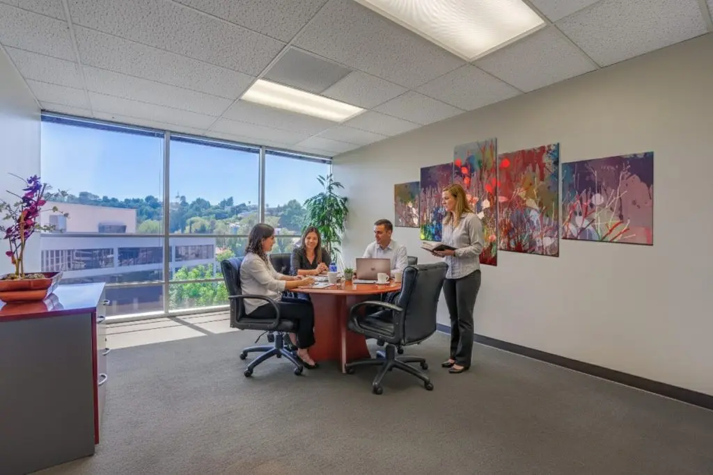 Barrister Suites Office Lease Four people are having a meeting in a modern office with large windows, colorful abstract art on the wall, and a round table with papers and coffee cups. One person stands while the others sit and talk.