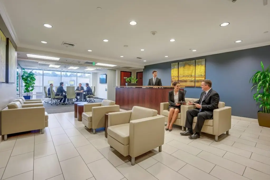 Barrister Suites Office Lease A modern office waiting area with beige chairs and plants. Two people in business attire sit talking, while others meet in a glass-walled conference room. A receptionist stands behind a wooden desk.