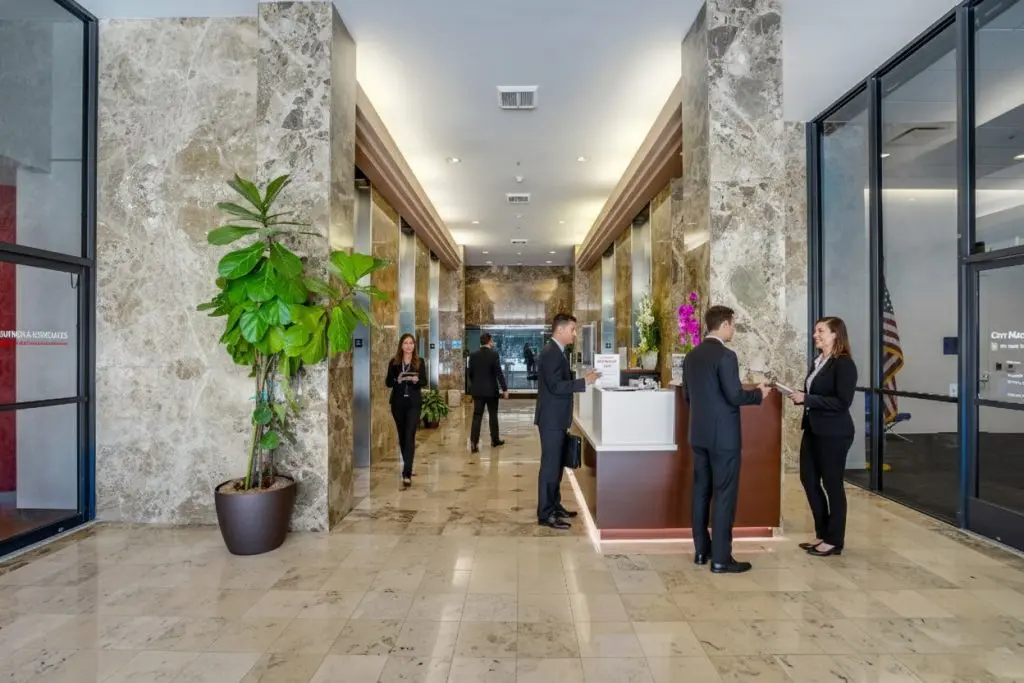 Barrister Suites Office Lease A modern office lobby with marble walls and floors, a large potted plant, and several people in business attire standing and talking near a reception desk.