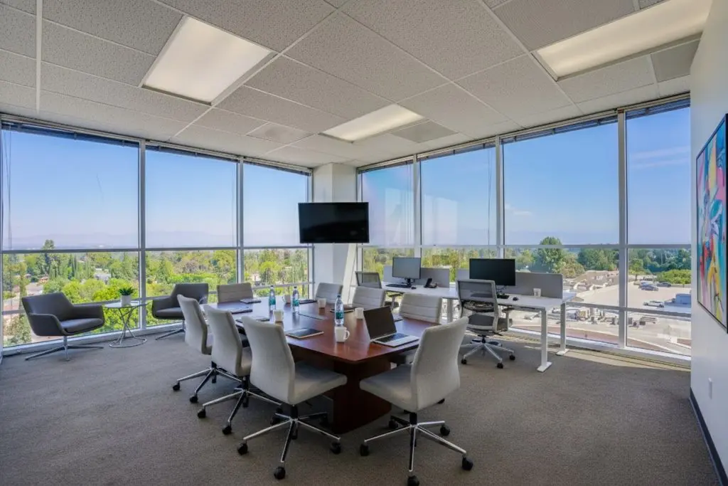 Barrister Suites Office Lease A modern office conference room with a large wooden table, gray chairs, laptops, monitors, and water bottles. Floor-to-ceiling windows provide a view of trees, a street, and blue sky outside. A TV is mounted on the wall.